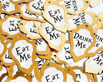 May include: A pile of gold-colored paper tags with a white background. The tags have a decorative border and the text "Eat Me" or "Drink Me" in black.