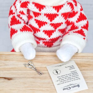 May include: A small elf doll wearing a red and white knitted sweater sits at a wooden table with a pair of silver scissors and a note that reads "Santa Claus Thank you so much for your order I hope it brings you and your miniature world joy Most of all! hope that you never lose that childlike sense of wonder and curiosity Sincerely Haddy The Hoonel Elf".