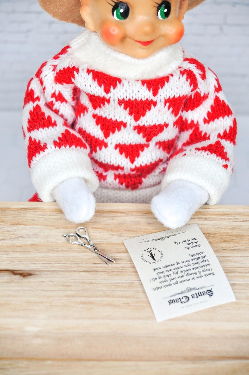 May include: A small elf doll wearing a red and white knitted sweater sits at a wooden table with a pair of silver scissors and a note that reads "Santa Claus Thank you so much for your order I hope it brings you and your miniature world joy Most of all! hope that you never lose that childlike sense of wonder and curiosity Sincerely Haddy The Hoonel Elf".