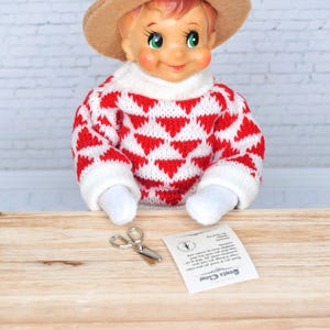 May include: A Christmas elf doll wearing a red and white sweater with a brown hat. The elf is holding a pair of small silver scissors and a note that says "Santa Claus".