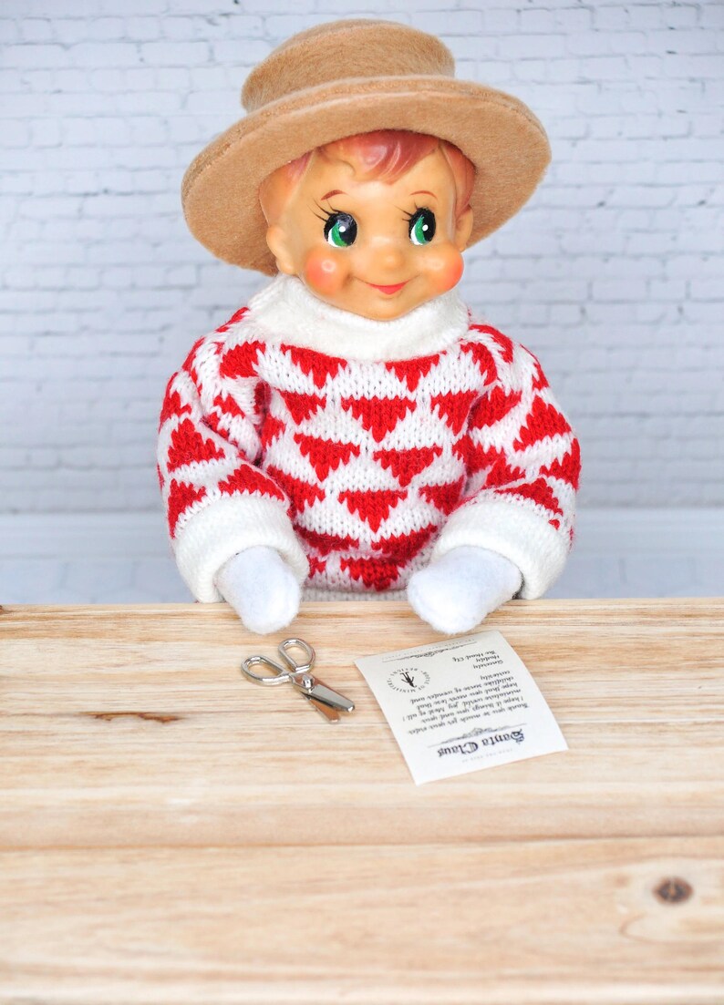 May include: A Christmas elf doll wearing a red and white sweater with a brown hat. The elf is holding a pair of small silver scissors and a note that says "Santa Claus".