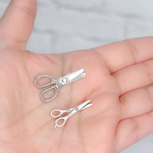 May include: Two silver-toned miniature scissors, one with a pointed tip and one with a rounded tip, are shown on a person's palm.
