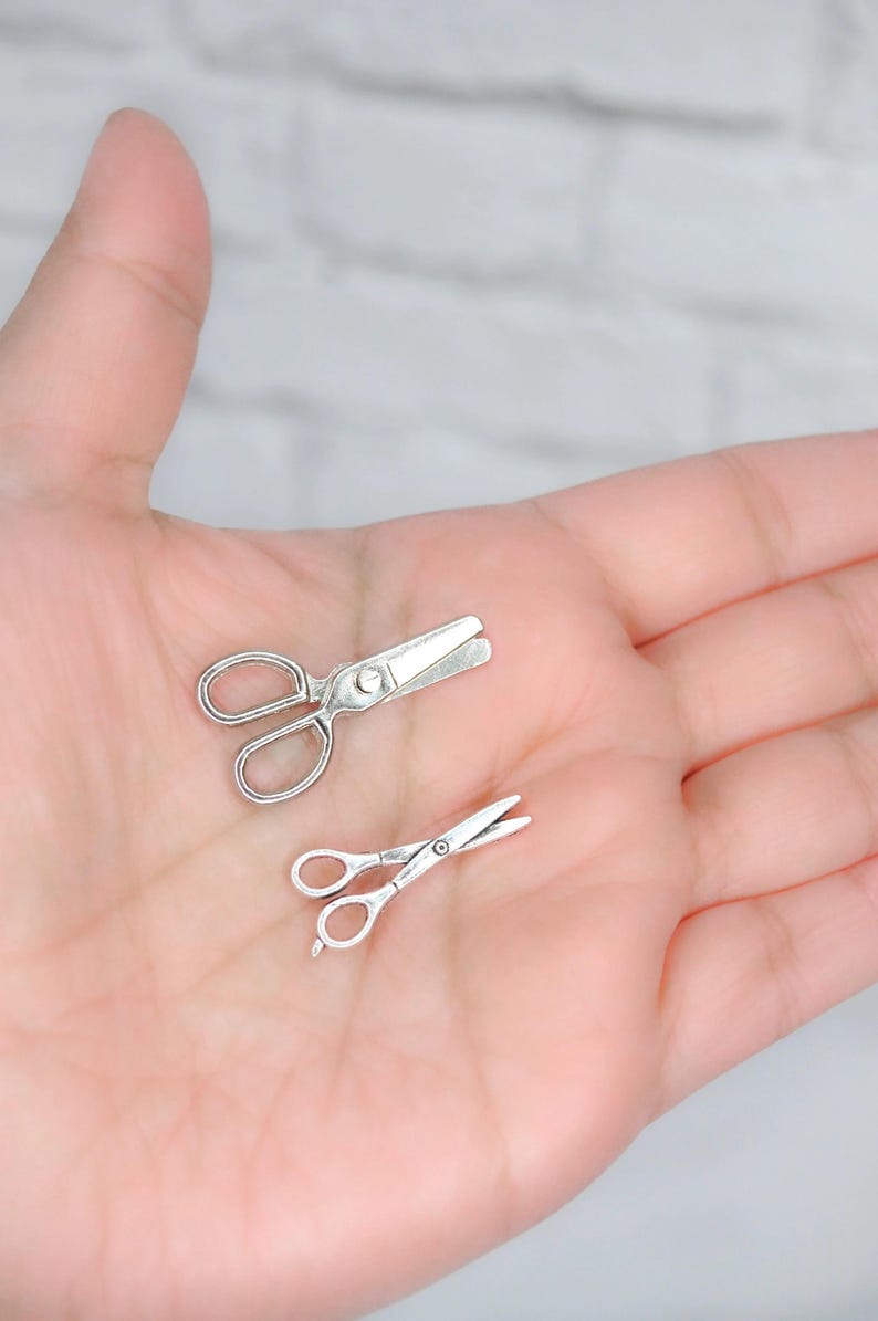 May include: Two silver-toned miniature scissors, one with a pointed tip and one with a rounded tip, are shown on a person's palm.