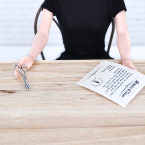 May include: A white card with black text that reads "Santa Claus Thank you so much for your order I hope it brings you and your miniature world joy Most of all I hope that you never lose that childlike sense of wonder The Hood Gardy"  A pair of silver scissors are on a wooden table.