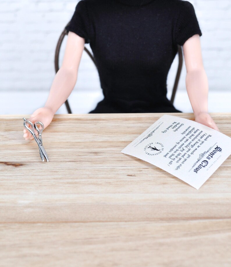 May include: A white card with black text that reads "Santa Claus Thank you so much for your order I hope it brings you and your miniature world joy Most of all I hope that you never lose that childlike sense of wonder The Hood Gardy"  A pair of silver scissors are on a wooden table.