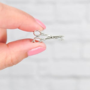 May include: A silver-toned pair of scissors charm, held between two fingers.