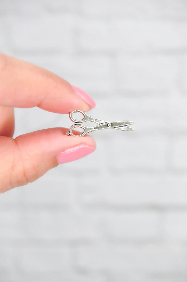 May include: A silver-toned pair of scissors charm, held between two fingers.