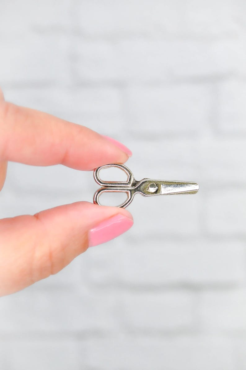 May include: A small, silver-colored pair of scissors held between two fingers.