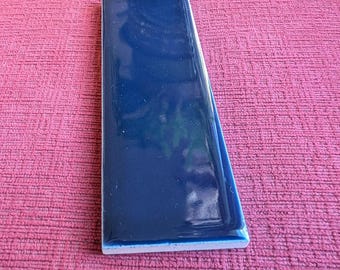 May include: A rectangular ceramic tile with a glossy, deep navy blue finish. The tile features a smooth, reflective surface and straight, clean edges, showcasing a uniform dark blue color throughout the entire top face of the piece.