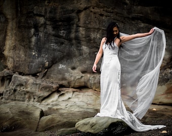 May include: A woman in a white wedding dress with a long veil blowing in the wind, standing on a rock formation.