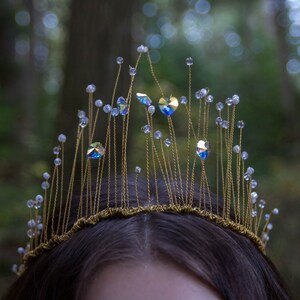 May include: A gold wire crown with iridescent blue crystals and white beads. The crown is worn on a person's head.