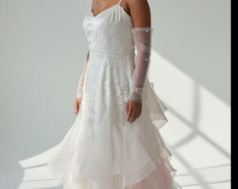 May include: A white floor-length gown with thin spaghetti straps, a V-neckline, and a bodice featuring intricate white floral embroidery. The skirt consists of layered, ruffled tulle with a subtle pink gradient at the hem, paired with sheer opera-length gloves adorned with small white pearls.