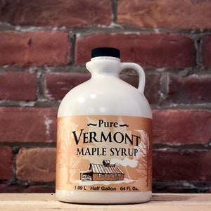 May include: A white ceramic jug with a black lid and a brown label that reads "Pure Vermont Maple Syrup" with a picture of a red barn and trees. The jug is 1.89 L, half gallon, and 64 fl. oz.