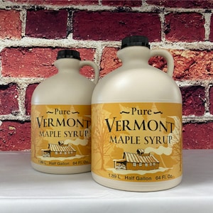 May include: Two plastic jugs of pure Vermont maple syrup. The jugs are labeled with the text "Pure Vermont Maple Syrup" and have a picture of a red barn with white trim. The jugs are 1.89 L or half gallon and contain 64 fl oz.