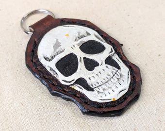 May include: A handmade leather keychain featuring a white skull design with black eye sockets and a stitched brown border. The keychain has a silver metal ring for attaching keys. The skull is painted on a dark brown leather base.