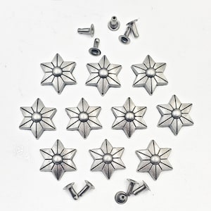 May include: A collection of silver-colored star-shaped metal studs and rivets. The star studs have a raised, detailed design with a central circular element. Several rivets are scattered around the stars, ready for application. The objects are arranged on a white surface.