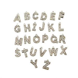 May include: A set of 26 silver letter charms, each letter is encrusted with clear rhinestones. The charms are arranged in alphabetical order.