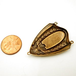 Puede incluir: Un hallazgo de joyer&iacute;a de metal de color bronce con un centro ovalado y patrones decorativos de hojas y flores. El hallazgo tiene tres peque&ntilde;os bucles para la fijaci&oacute;n. Una moneda estadounidense est&aacute; al lado para la comparaci&oacute;n de tama&ntilde;o.