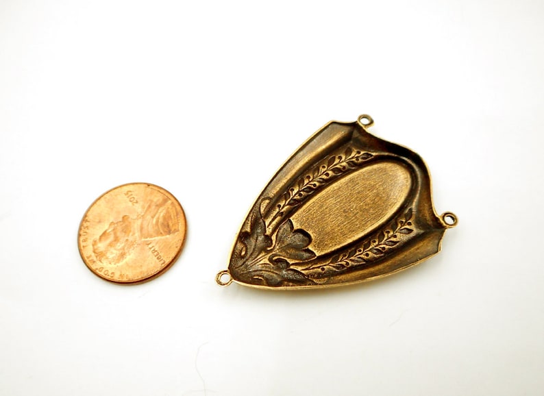 Puede incluir: Un hallazgo de joyer&iacute;a de metal de color bronce con un centro ovalado y patrones decorativos de hojas y flores. El hallazgo tiene tres peque&ntilde;os bucles para la fijaci&oacute;n. Una moneda estadounidense est&aacute; al lado para la comparaci&oacute;n de tama&ntilde;o.