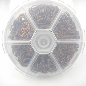 May include: A clear, segmented container filled with numerous small, bronze-colored jump rings. The container is round with a hexagonal center and is set against a white background. The jump rings are tightly packed, suggesting a large quantity.