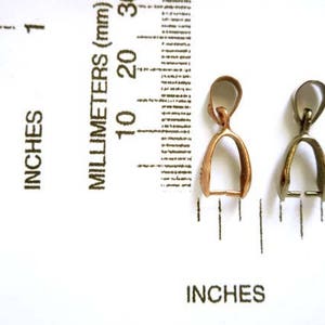 May include: Two metal pendant bails, one gold and one silver, are shown next to a ruler. The bails are about 1 inch long.