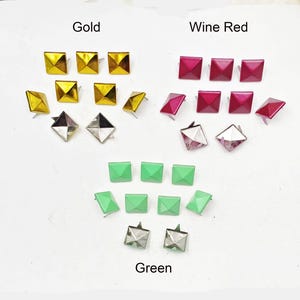 May include: Assortment of pyramid-shaped metal studs in gold, wine red, and green colors. The studs are arranged in rows on a white surface.