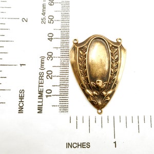 Puede incluir: Un colgante de escudo de metal color bronce con un centro ovalado y un dise&ntilde;o floral y de hojas. El colgante tiene dos peque&ntilde;os agujeros en la parte superior y uno en la inferior. La imagen incluye una regla con pulgadas y mil&iacute;metros.