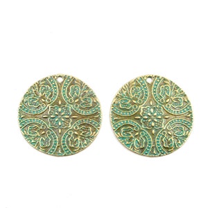 以下が含まれることがあります： 緑と金の複雑な花柄が施された丸い金属製のチャームが2つ。チャームの上部には、ジュエリーやその他のクラフトに取り付けるための穴があります。