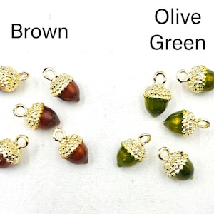 May include: A collection of acorn-shaped charms featuring smooth brown or olive green bodies and textured gold-tone metal caps. Each charm includes a small circular loop at the top for attachment. The text Brown and Olive Green labels the groups.