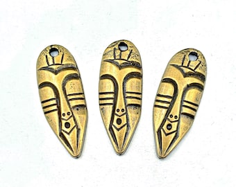 May include: Three antique gold-colored tribal mask pendants. Each pendant features a stylized face with horizontal lines, a diamond-shaped mouth, and a hole at the top for stringing. The pendants have a weathered, aged appearance.
