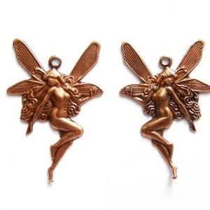 Op de afbeelding: Twee koperkleurige fee-bedels met vleugels en golvend haar. De feeën zijn in een vliegende pose met uitgestrekte armen.