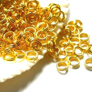 May include: A pile of gold-coloured double jump rings, scattered on a white, textured surface. The rings are small, with a shiny finish, and appear to be made of metal. The image is well-lit, highlighting the details of the jewellery findings.