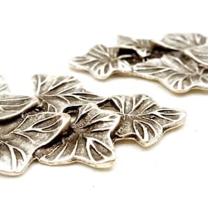 May include: Silver-toned metal leaf-shaped charms with a textured surface. The charms are arranged in a cluster, with the largest charm in the center.