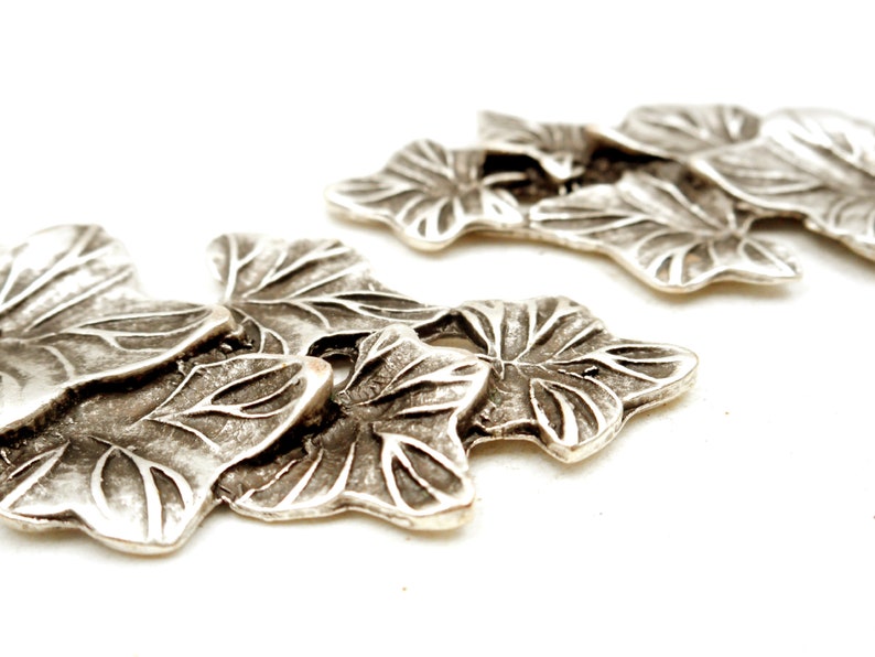 May include: Silver-toned metal leaf-shaped charms with a textured surface. The charms are arranged in a cluster, with the largest charm in the center.