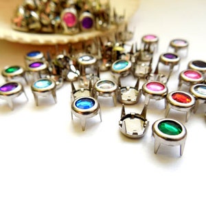 Puede incluir: Una colección de tachuelas de metal plateado con centros de gemas facetadas de colores. Las gemas son de varios colores, incluyendo azul, verde, rosa, morado y rojo. Las tachuelas tienen tres pequeñas patas y están dispersas sobre una superficie blanca.