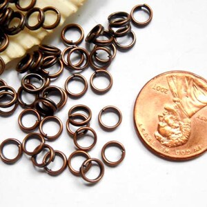 May include: Close-up of small, bronze-colored jump rings scattered on a white surface. A U.S. penny from 2007 is next to the rings for size comparison. The rings are open and ready for use in jewelry making or crafts.