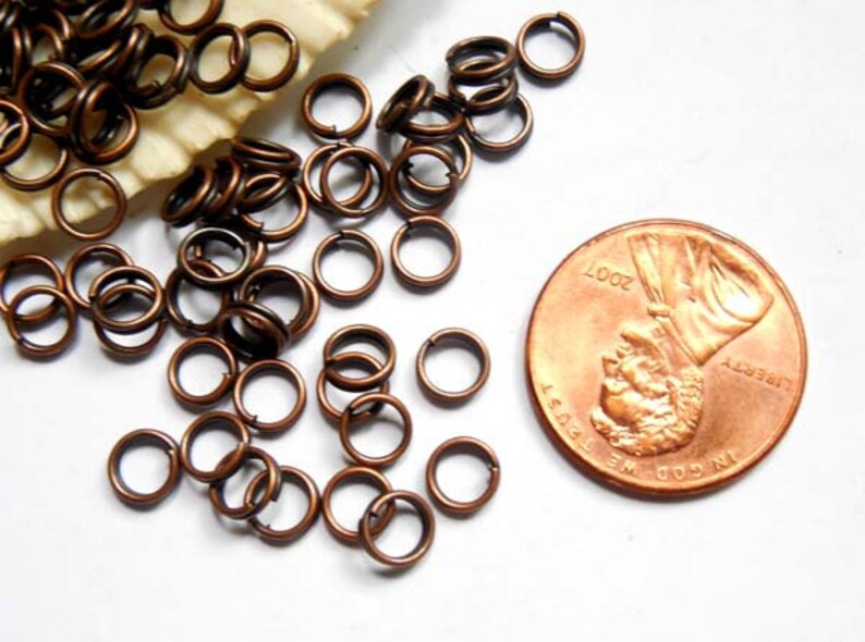 May include: Close-up of small, bronze-colored jump rings scattered on a white surface. A U.S. penny from 2007 is next to the rings for size comparison. The rings are open and ready for use in jewelry making or crafts.