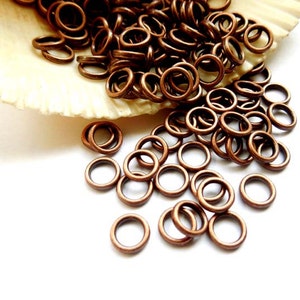 May include: A large pile of small, copper-coloured jump rings. The rings are scattered on a white surface and a seashell.