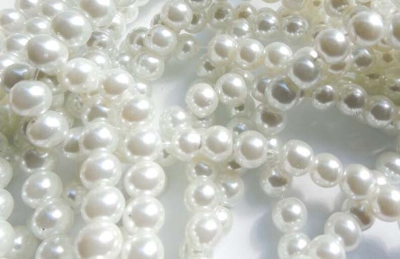 May include: A close-up of a pile of white pearl beads. The beads are round and smooth, and they are arranged in a random pattern.
