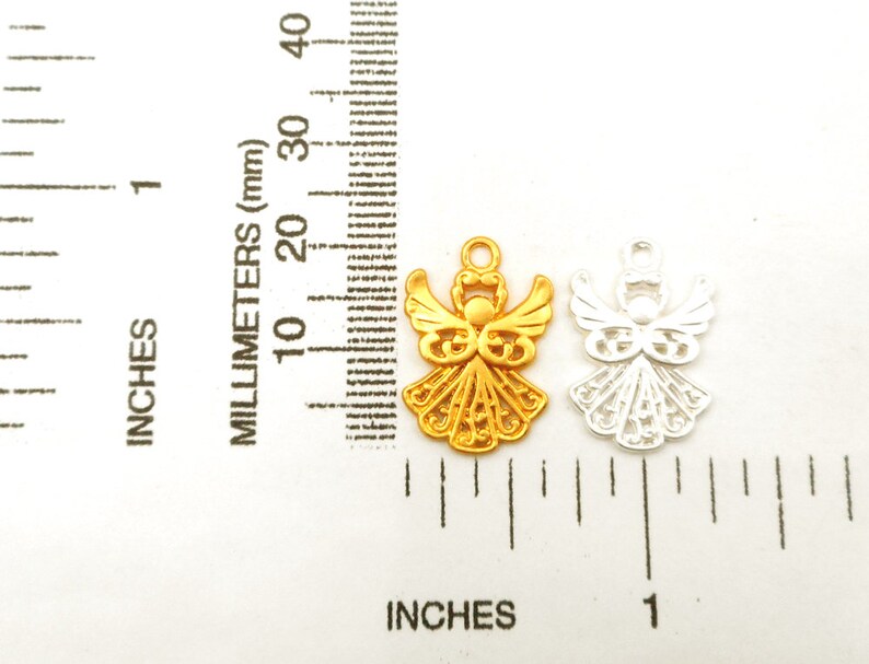 May include: Two angel charms, one gold and one silver, with intricate designs. The charms are about 1 inch tall and are shown next to a ruler.