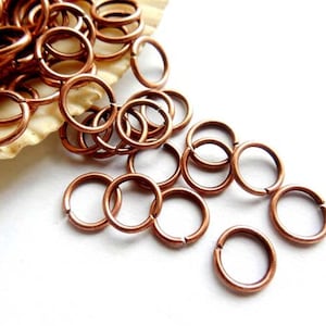 May include: A pile of copper jump rings, some are scattered on a white surface. The jump rings are small and round, and they are perfect for jewelry making.