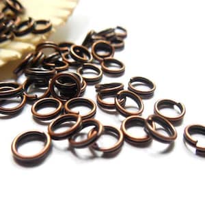 May include: Close-up of a collection of copper-colored double split rings, scattered on a white surface. The rings are made of metal and have a dark inner core. These jewelry findings are used for connecting jewelry components.