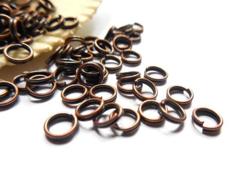 May include: Close-up of a collection of copper-colored double split rings, scattered on a white surface. The rings are made of metal and have a dark inner core. These jewelry findings are used for connecting jewelry components.