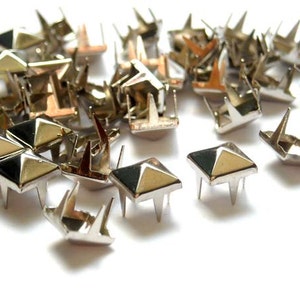May include: A close-up of a pile of silver pyramid-shaped studs with three prongs on the bottom. The studs are scattered on a white surface.