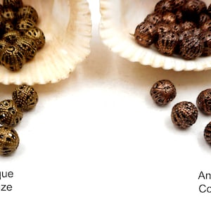 May include: Two piles of decorative metal beads, one in antique bronze and one in antique copper. The beads are round and have a delicate, intricate design.