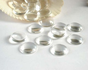 May include: A group of clear glass cabochons, also known as glass domes, are scattered on a white surface. The cabochons are round and have a smooth, polished finish.