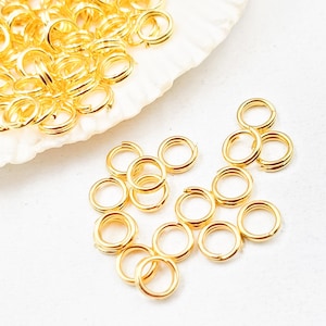 May include: A collection of small, shiny gold-colored double jump rings. These jewelry-making components are arranged on a white surface, with some rings clustered together and others scattered. The rings have a smooth, polished finish.