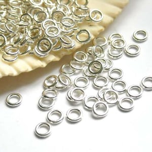 May include: A large pile of silver jump rings, scattered on a white surface. The jump rings are small and round, with a smooth finish.