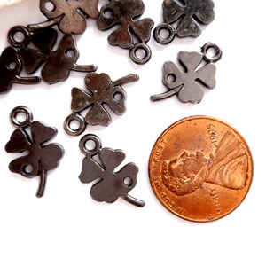 May include: A close-up of a group of black metal four-leaf clover charms with a jump ring. The charms are scattered on a white surface next to a US penny.