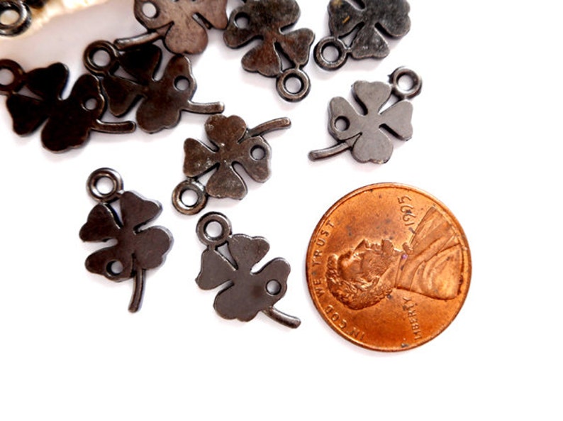 May include: A close-up of a group of black metal four-leaf clover charms with a jump ring. The charms are scattered on a white surface next to a US penny.
